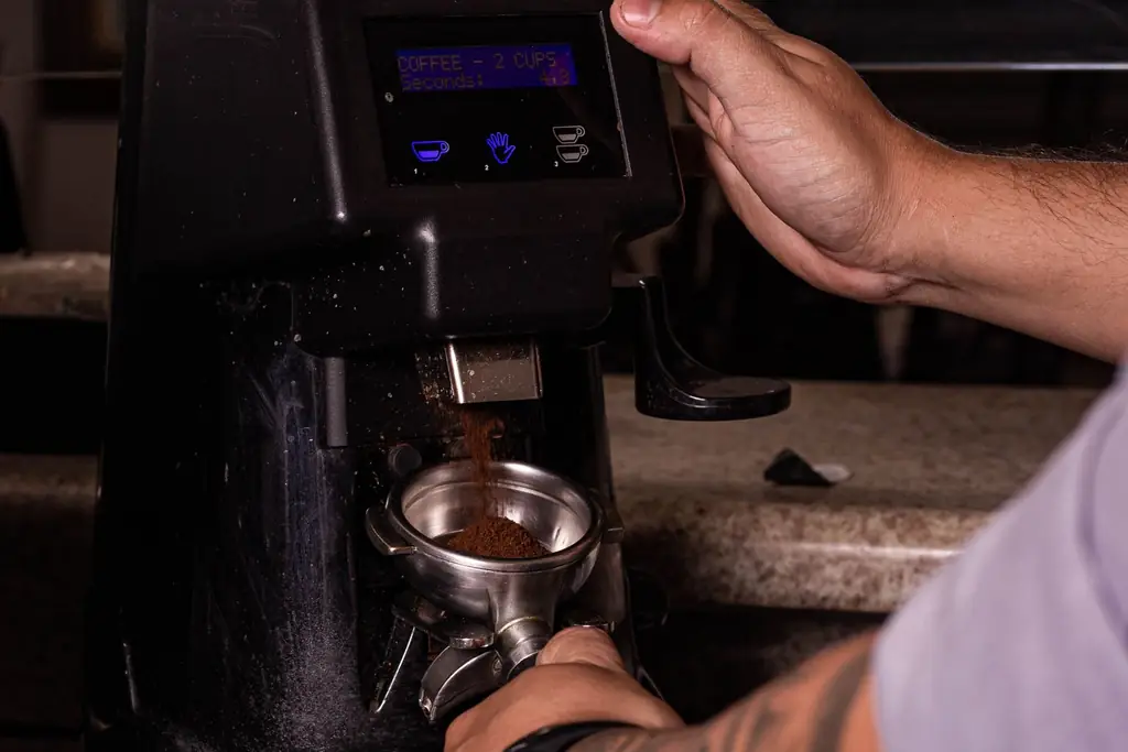Barista holding a portafilter under a black electric espresso grinder (illustrative, not a specific model)