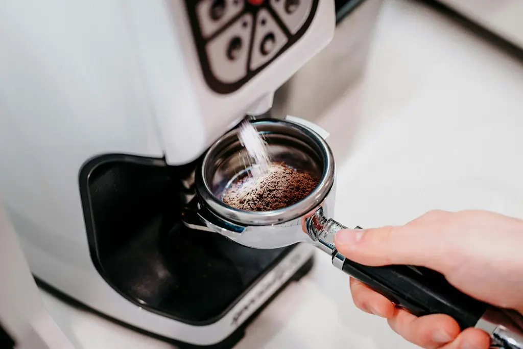 Hand holding a portafilter under a white espresso grinder as grounds fall